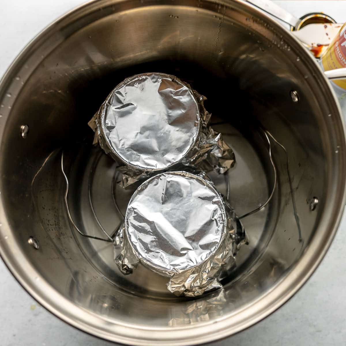 Two cans covered with aluminum foil sit on a rack inside a large stainless steel pot, prepared for a water bath.