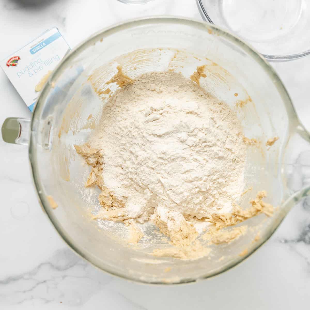 A clear mixing bowl contains dough and a pile of white flour.
