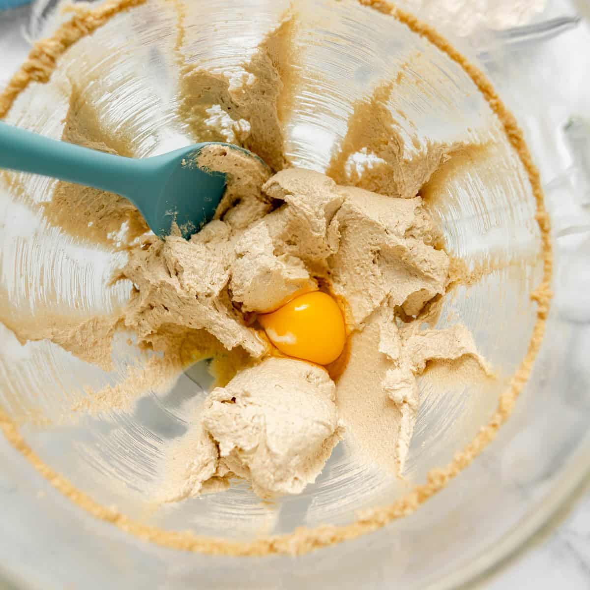 A blue spatula in a glass bowl with creamed butter and sugar mixture and a cracked raw egg yolk in the center.