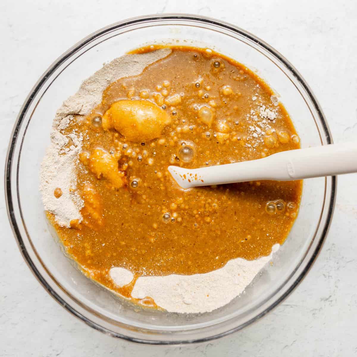 A glass bowl containing a mixture of flour and wet ingredients with a white spatula resting inside, ready for mixing.