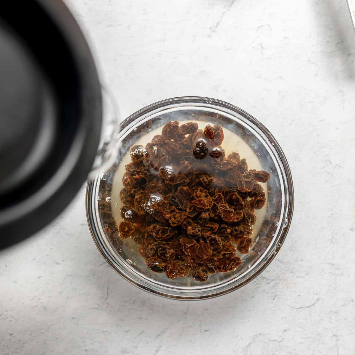 A glass bowl filled with raisins soaking in water on a light-colored surface.