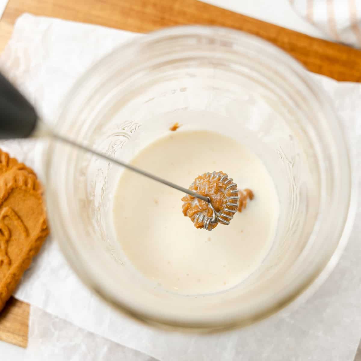 A handheld milk frother blends a brown ingredient into milk inside a glass jar, with a cookie and parchment paper on a wooden board nearby.