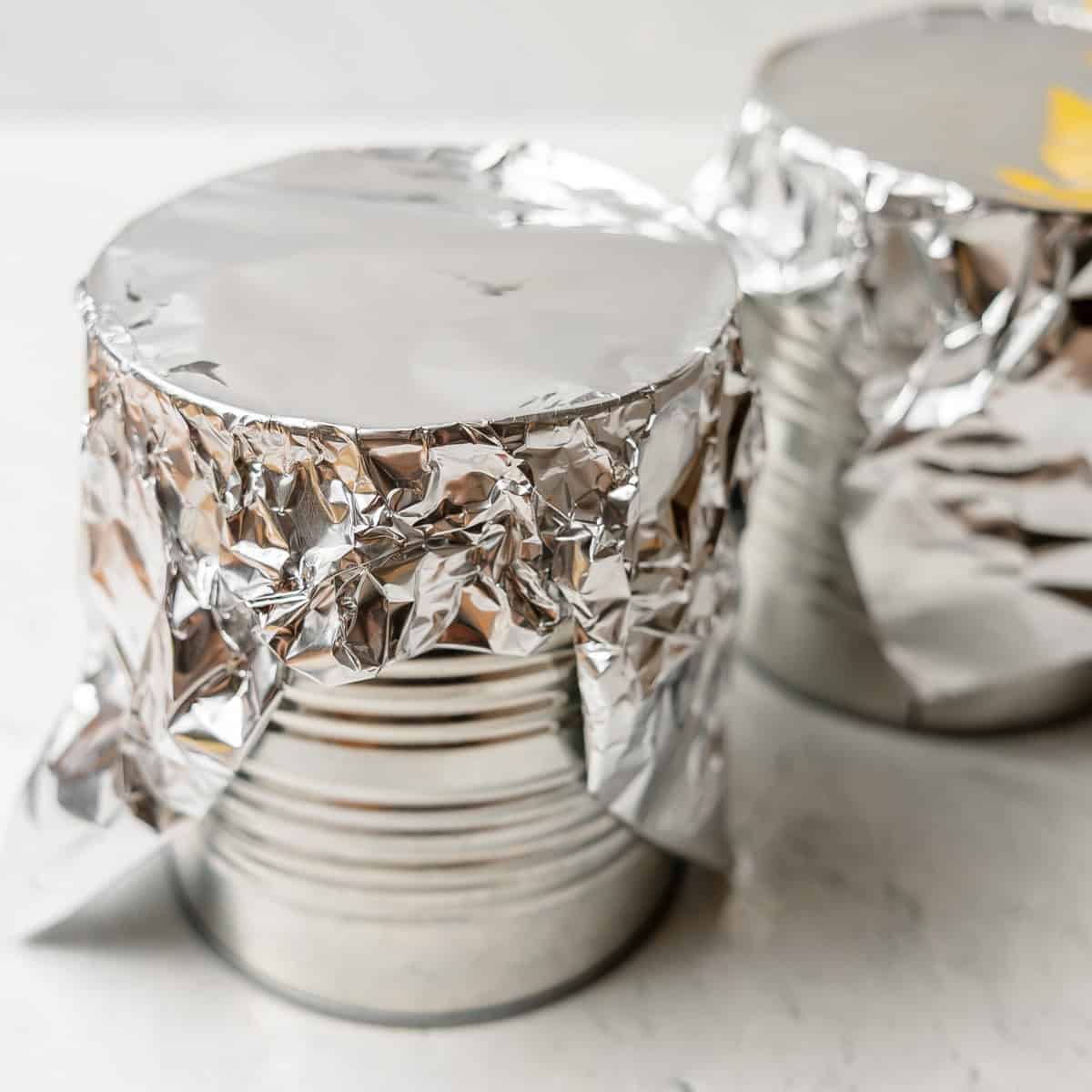 Two metal cans are covered with aluminum foil, sitting on a white surface.