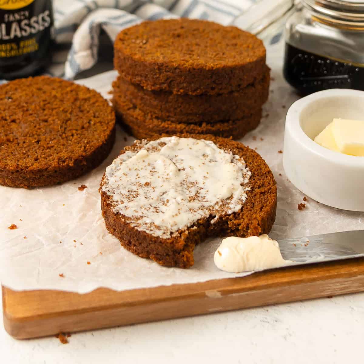 Slices of dark bread on parchment paper, with one slice spread with butter and a bite taken out. A butter knife, a small dish of butter, and a jar are visible nearby.