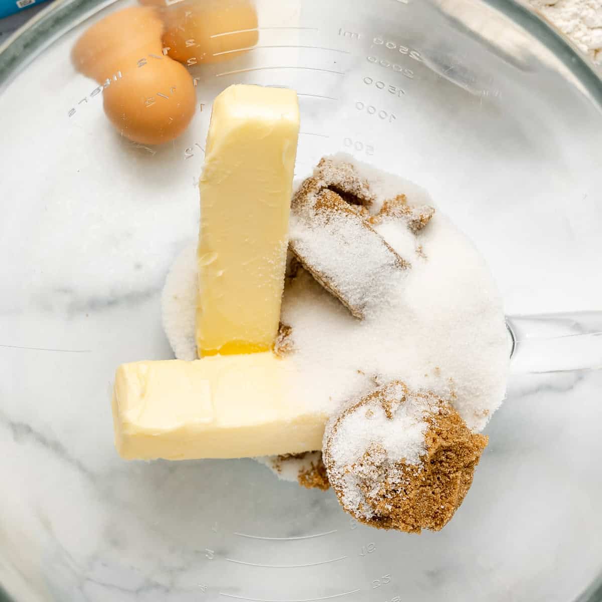 Two sticks of butter, granulated sugar, brown sugar, and eggs in a glass mixing bowl on a marble surface.
