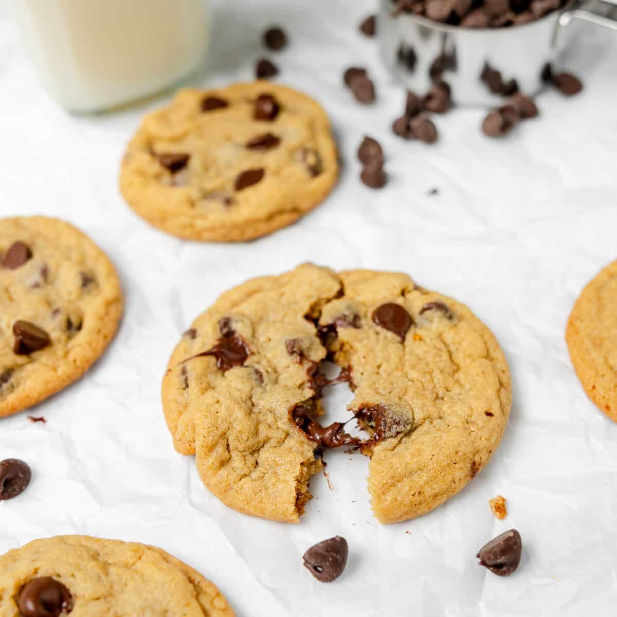 A chocolate chip cookie broken in half sits on crumpled parchment paper, surrounded by whole cookies and scattered chocolate chips.