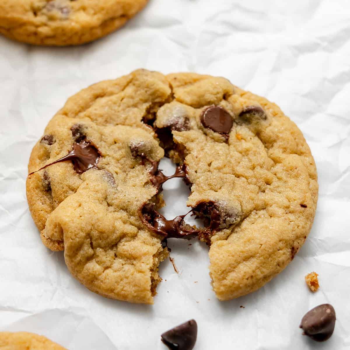 A chocolate chip cookie broken in half on white parchment paper, with melted chocolate chips visible inside.