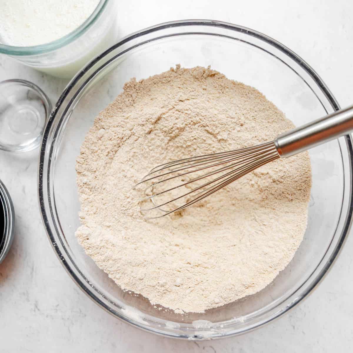 A metal whisk in a glass bowl of flour mixture on a white surface, with a glass measuring cup and small bowl nearby.