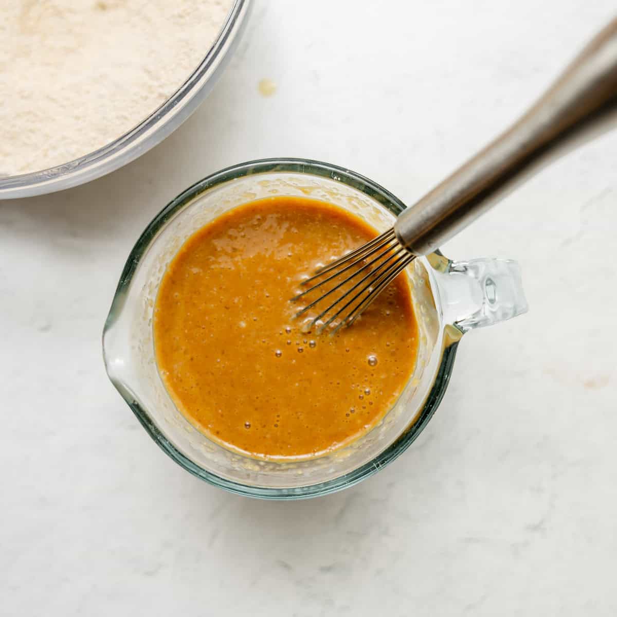 A metal whisk rests in a glass measuring cup filled with a smooth brown batter, with a bowl of dry ingredients visible in the background.