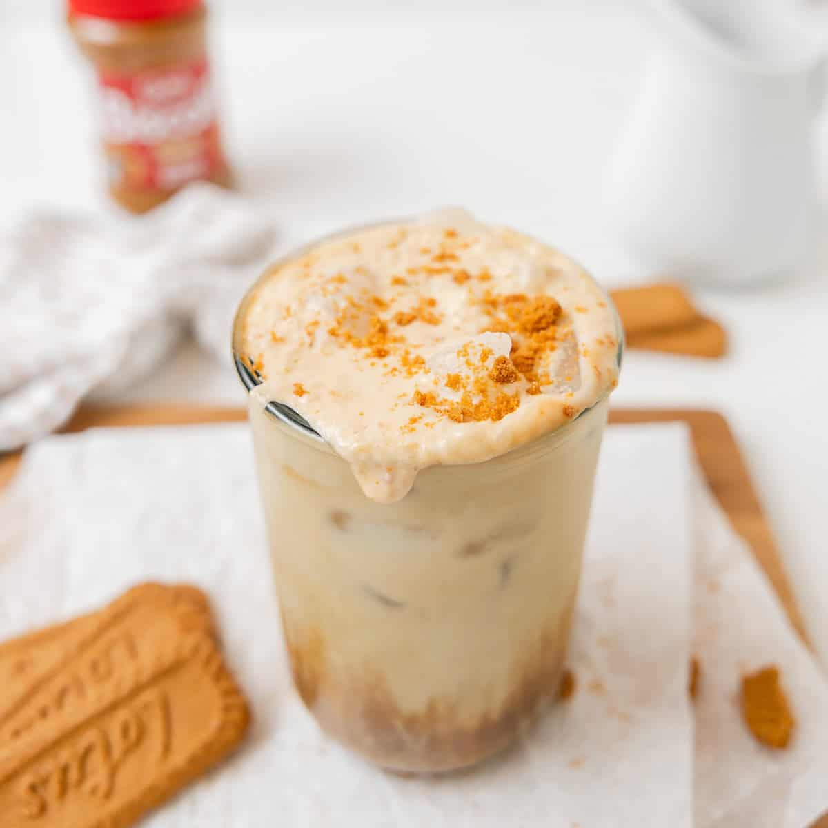 A glass of iced coffee topped with whipped cream and cookie crumbs, with a cookie and a jar of cookie butter in the background.