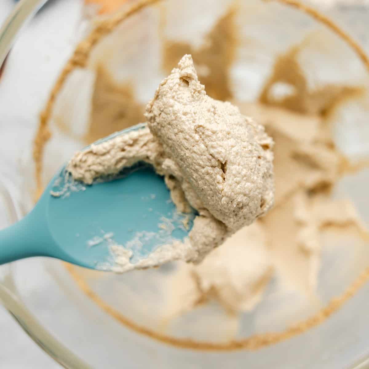 A close-up of a blue spatula holding a scoop of thick, beige batter over a glass mixing bowl.