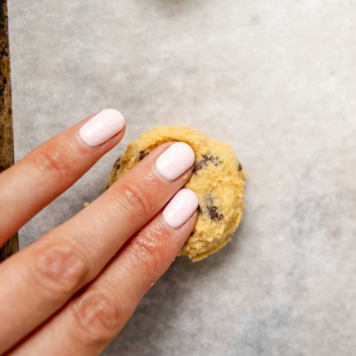 A hand with pale pink nail polish presses down on a round ball of chocolate chip cookie dough on a parchment-lined baking sheet.