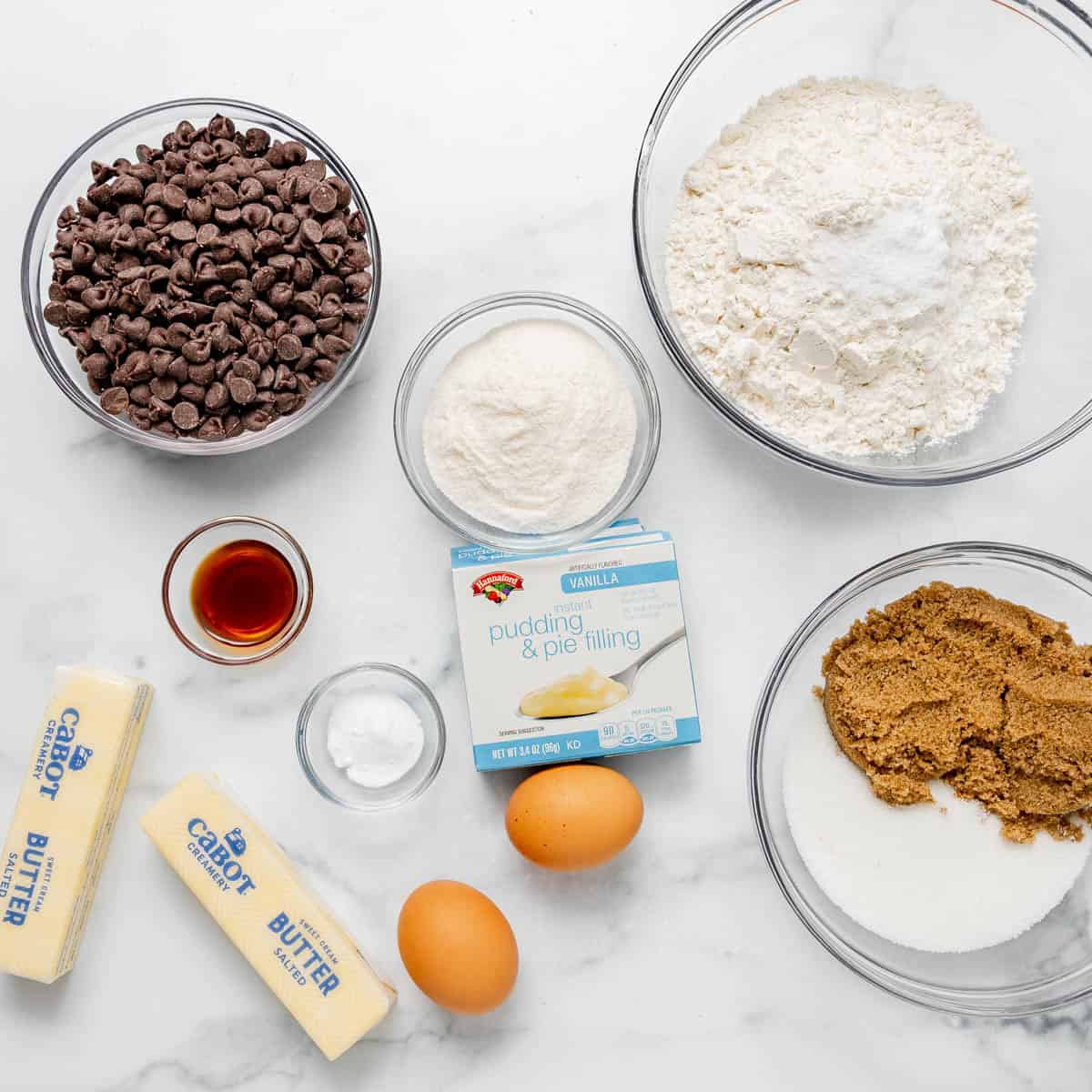 Baking ingredients on a white surface include chocolate chips, flour, brown sugar, white sugar, vanilla extract, baking soda, salt, two eggs, butter, and vanilla pudding mix.