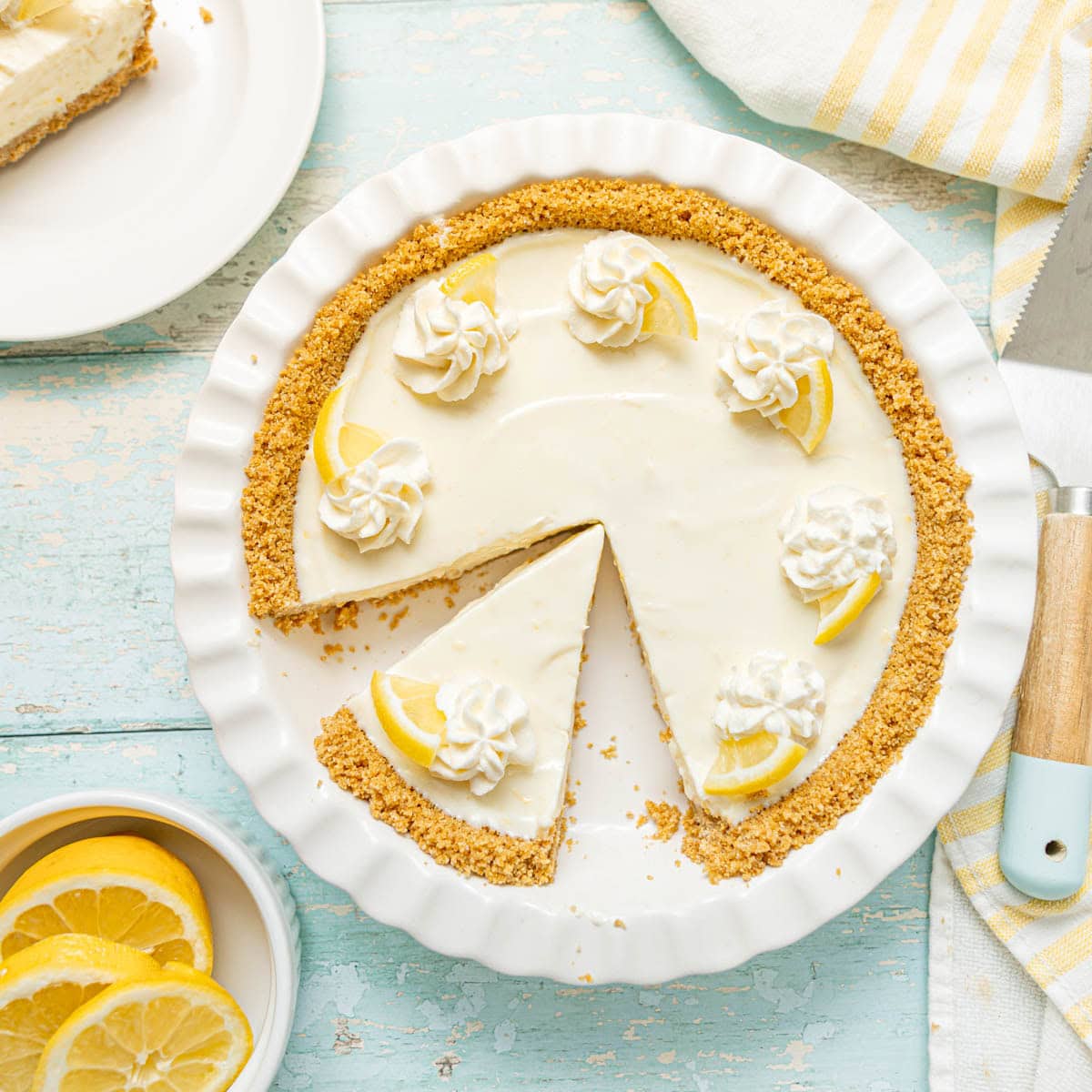 A lemon cream pie with a graham cracker crust, decorated with whipped cream and lemon slices, with one slice removed and placed on a plate.