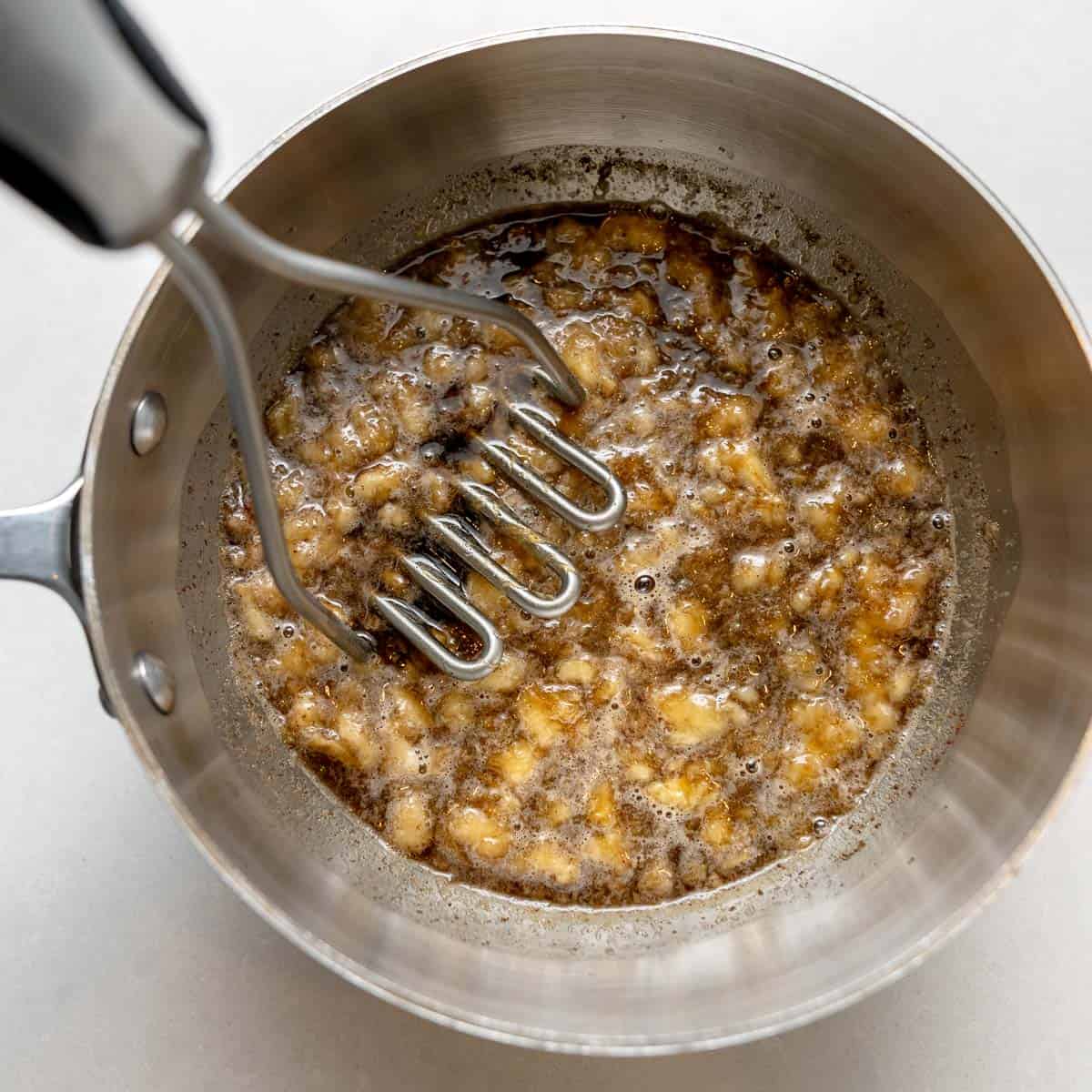 A potato masher crushing bananas in a saucepan, with the mixture appearing browned and partially mashed.