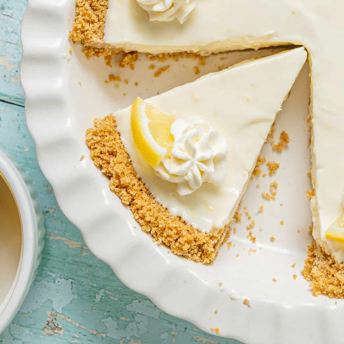 A lemon cream pie with a graham cracker crust in a white dish, with one slice missing and lemon wedge garnish, next to a cup of coffee on a blue wooden surface.