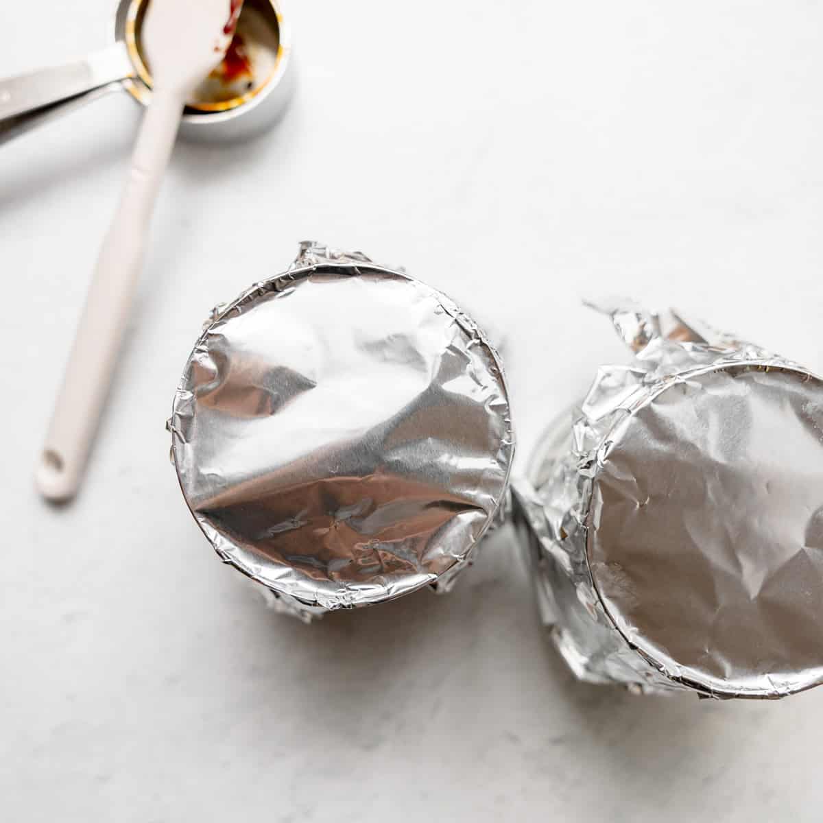 Two cans covered with aluminum foil on a light surface, next to a measuring spoon and a spatula.