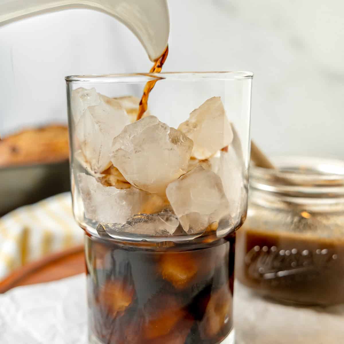 A glass filled with ice cubes being poured with cold brew coffee, with a jar of coffee concentrate in the background.