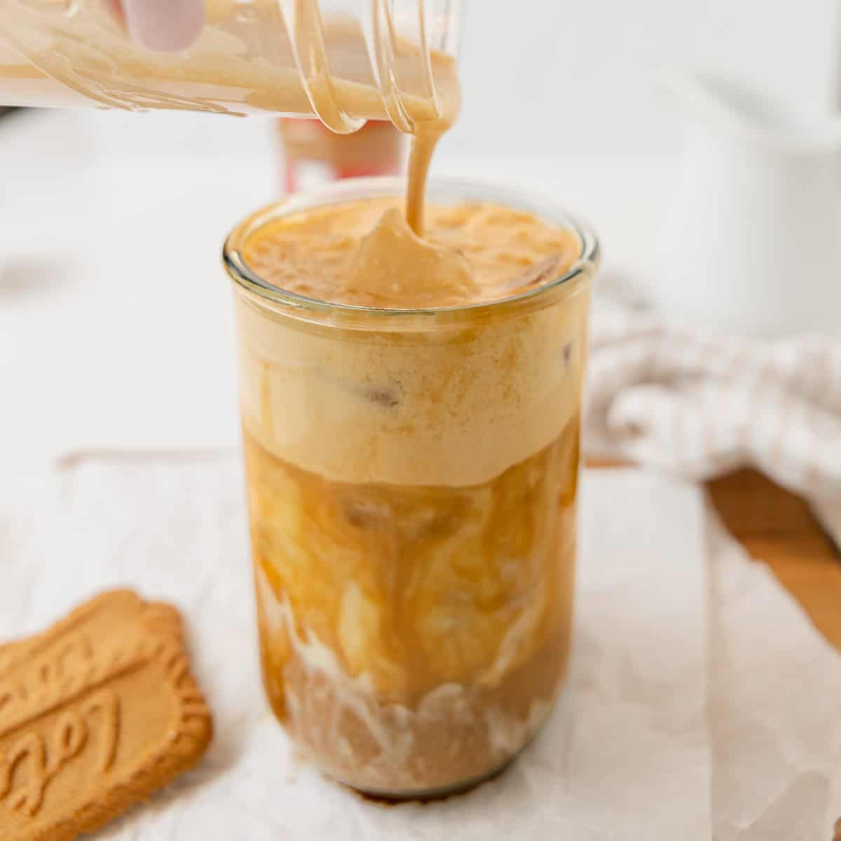 A creamy mixture is being poured into a glass of iced coffee, creating a swirling effect. A cookie lies on the side, and a light-colored cloth is visible in the background.