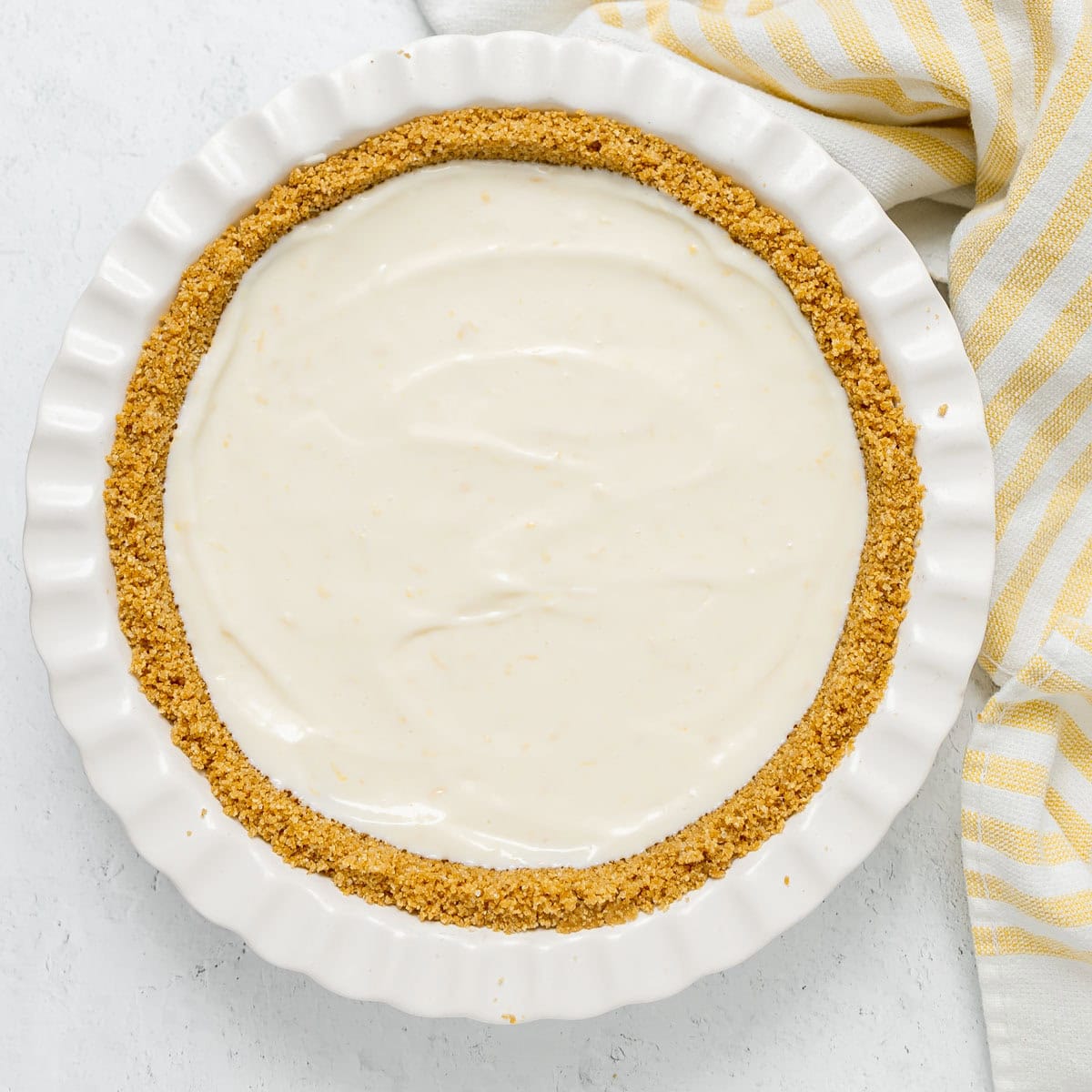 A pie dish with a graham cracker crust filled with a smooth, creamy white filling sits next to a yellow and white striped cloth.
