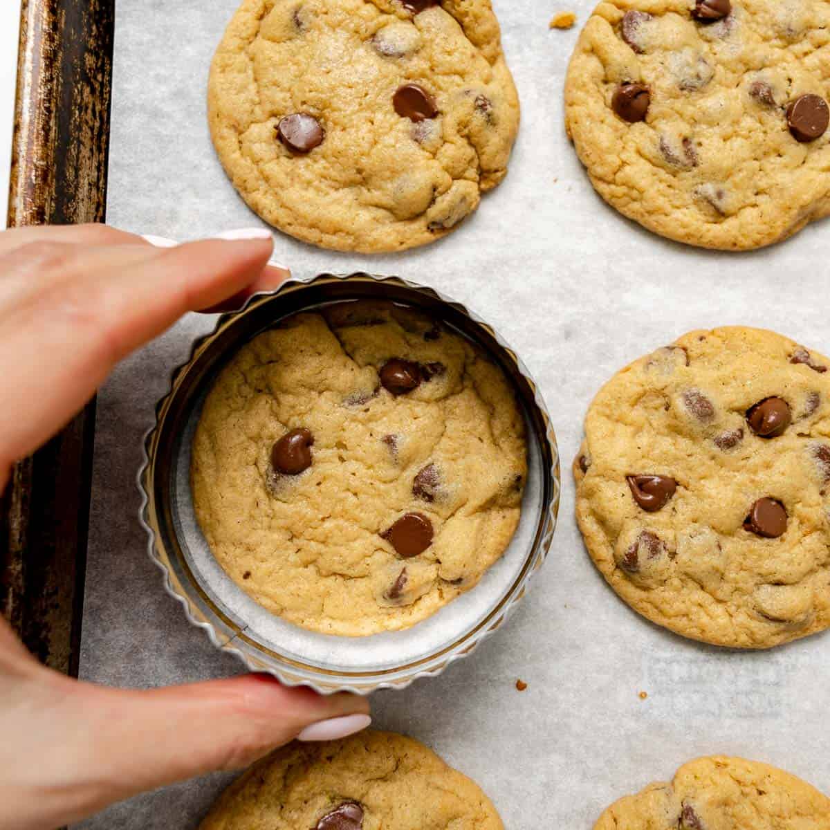 A hand holds a metal cookie cutter over a freshly baked chocolate chip cookie on a baking sheet with other cookies.