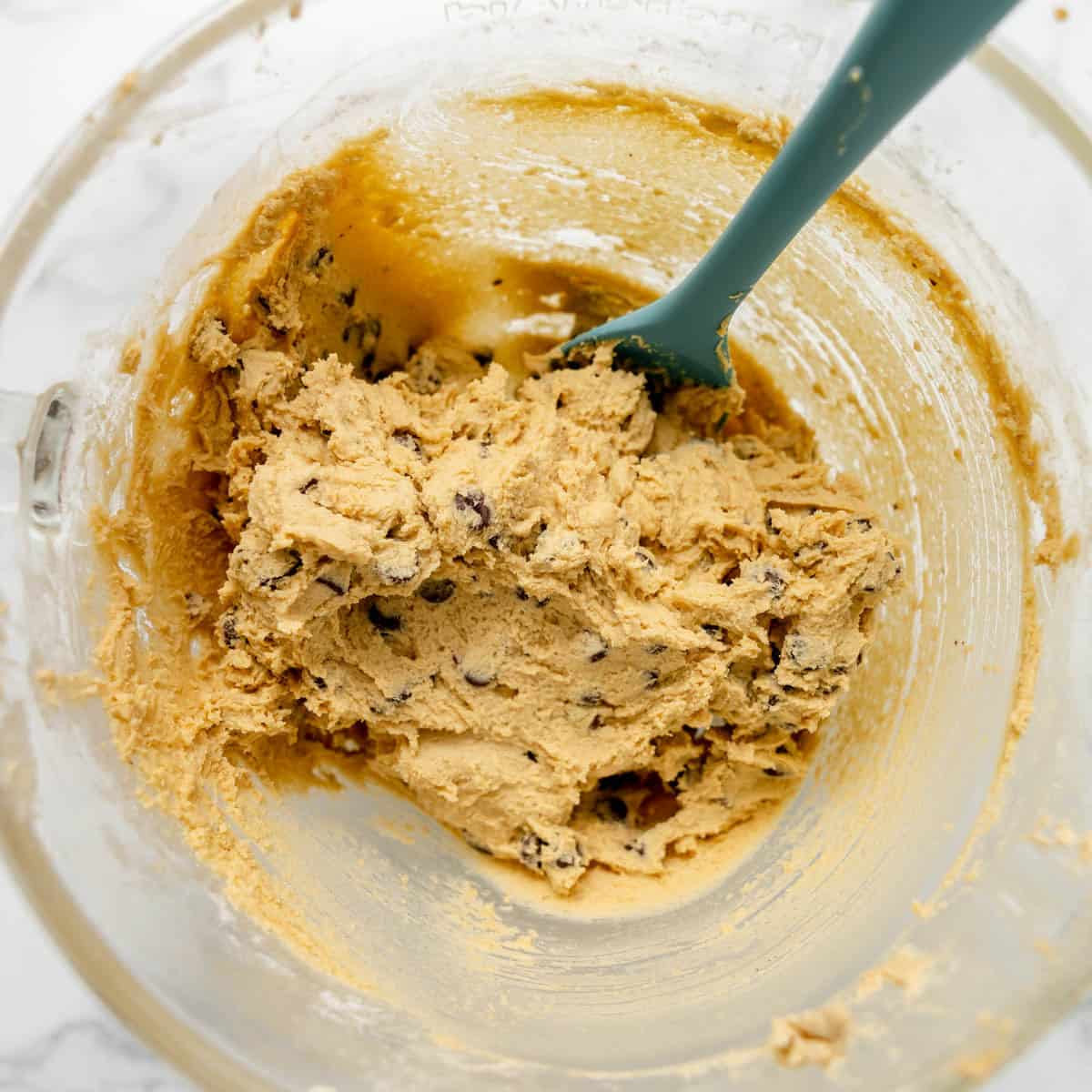 A glass mixing bowl containing chocolate chip cookie dough with a green spatula resting inside.