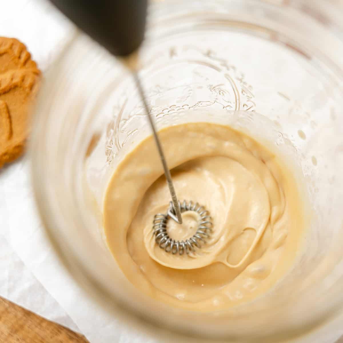 A handheld milk frother mixes a creamy, light brown liquid in a glass jar, viewed from above.