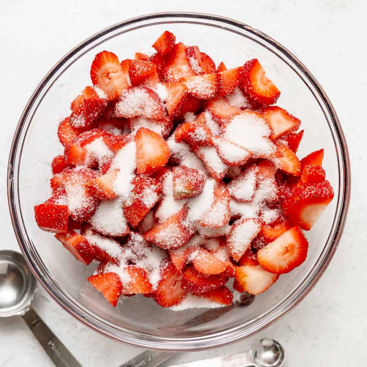 A glass bowl filled with sliced strawberries topped with granulated sugar, with measuring spoons placed beside the bowl on a white surface.