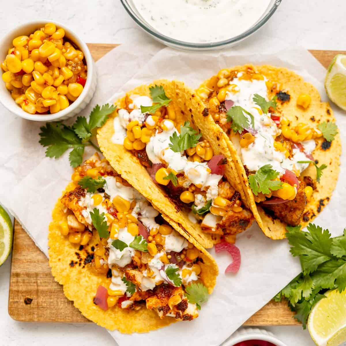 Three tacos filled with seasoned fish, corn, diced vegetables, and cilantro, topped with a creamy sauce, served on a wooden board with lime wedges and a side of corn.