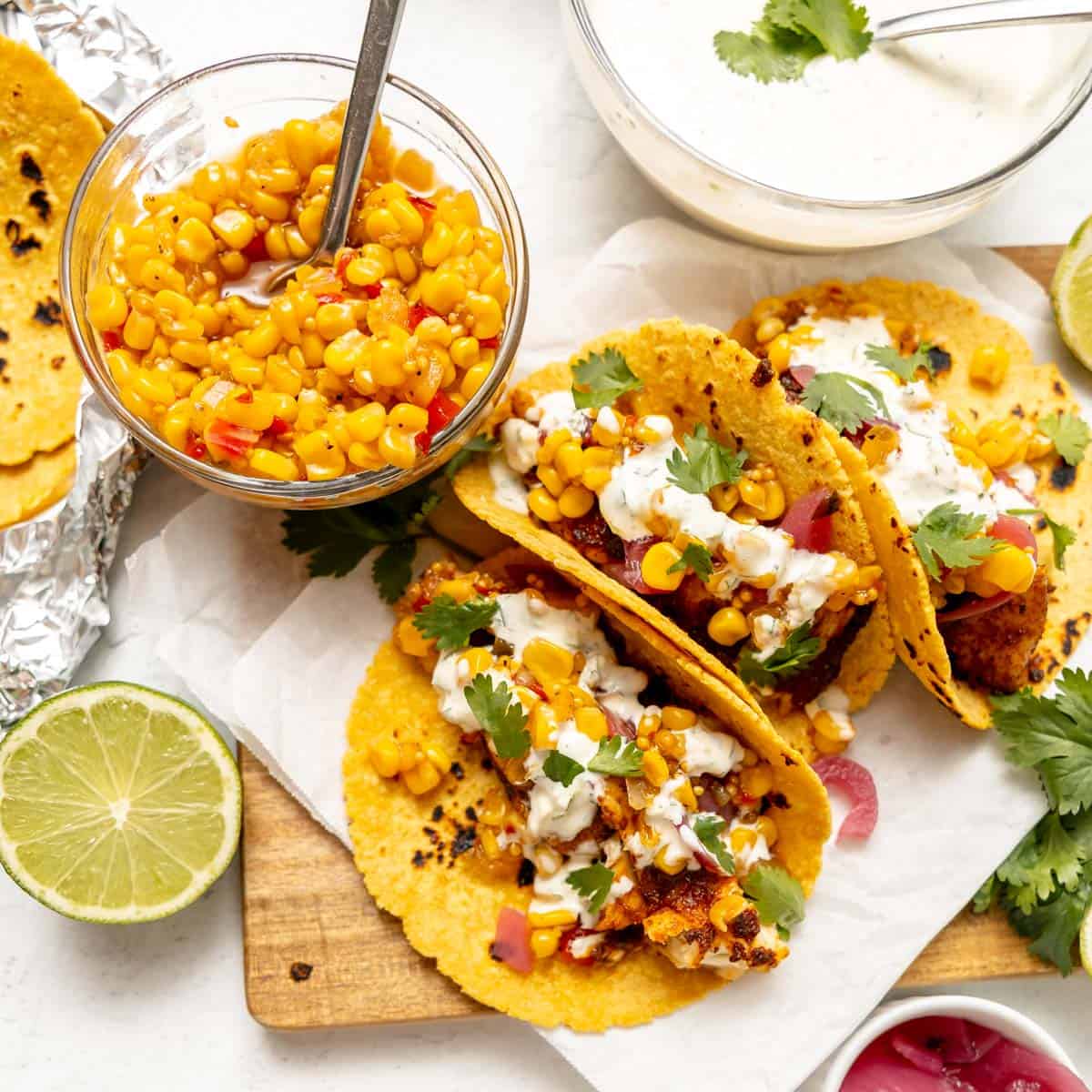 Three corn tortillas filled with seasoned fish, corn salsa, white sauce, and cilantro are arranged on a plate with lime, a bowl of corn salsa, and fresh cilantro on the side.