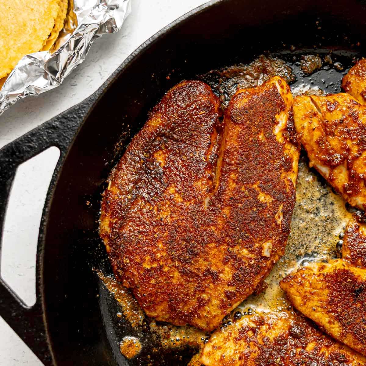 Cast iron skillet with blackened, seasoned fish fillets being cooked, next to a stack of foil-wrapped tortillas on a white surface.