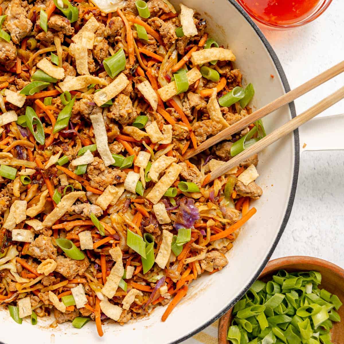 A skillet filled with an egg roll in a bowl mixture featuring ground meat, shredded carrots, cabbage, crispy wonton strips, and chopped green onions, with chopsticks on the side.