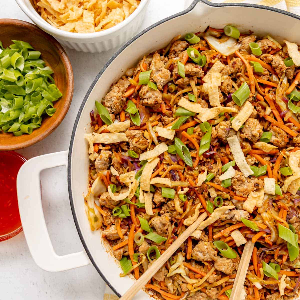 A skillet filled with an egg roll in a bowl dish, containing ground meat, shredded vegetables, and green onions, with bowls of green onions, sauce, and crispy strips on the side.