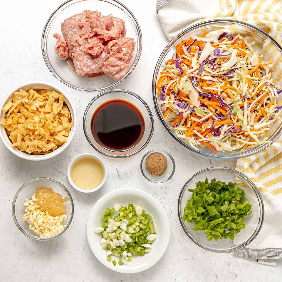 Ingredients for an Asian-inspired dish laid out in bowls, including ground meat, coleslaw mix, soy sauce, green onions, garlic, ginger, seasonings, and crispy noodles.