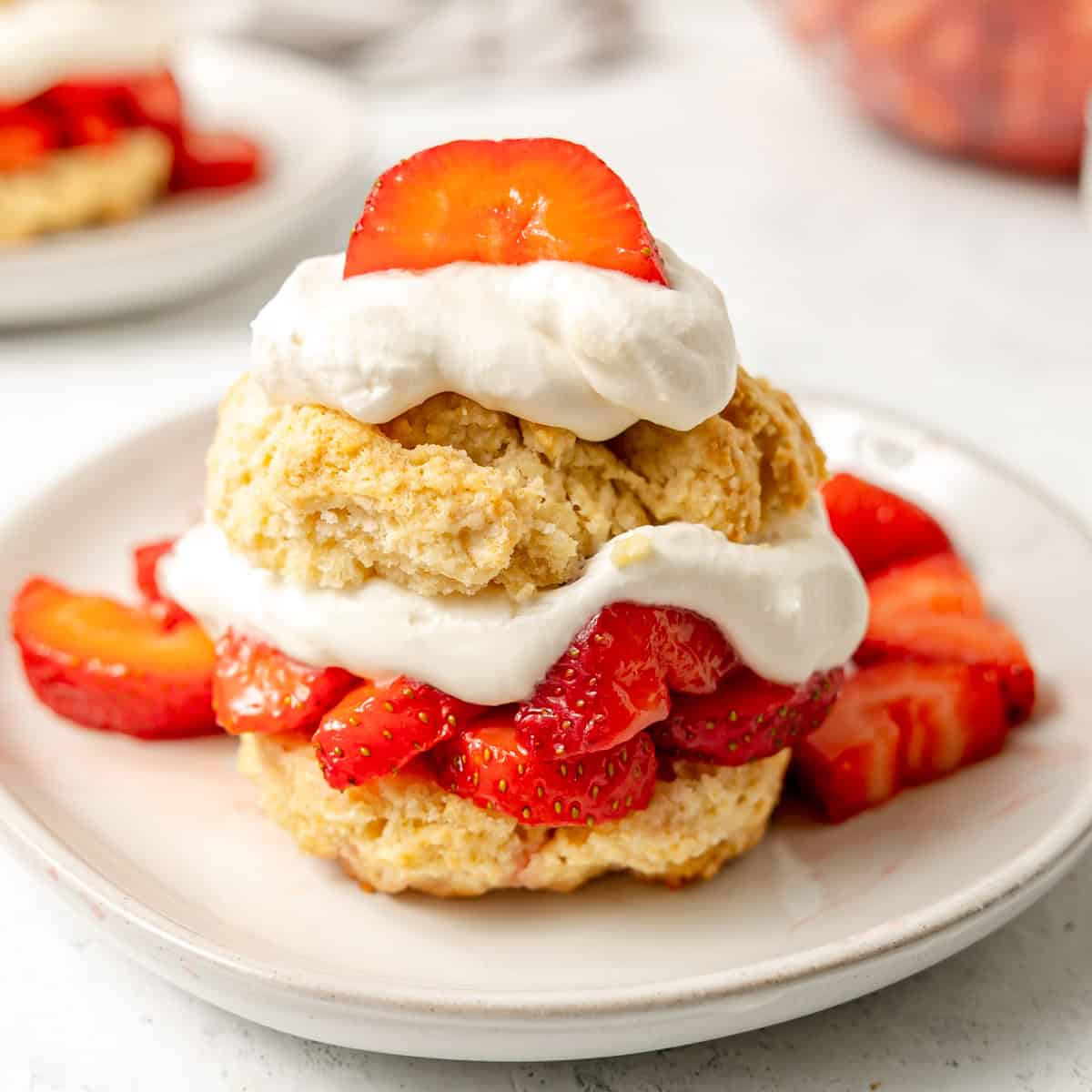 A strawberry shortcake dessert topped with whipped cream and fresh sliced strawberries on a white plate.