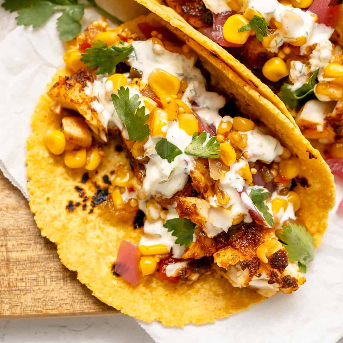 Two corn tortillas filled with grilled chicken, corn, red onion, cilantro, and a creamy white sauce, arranged on a wooden board.