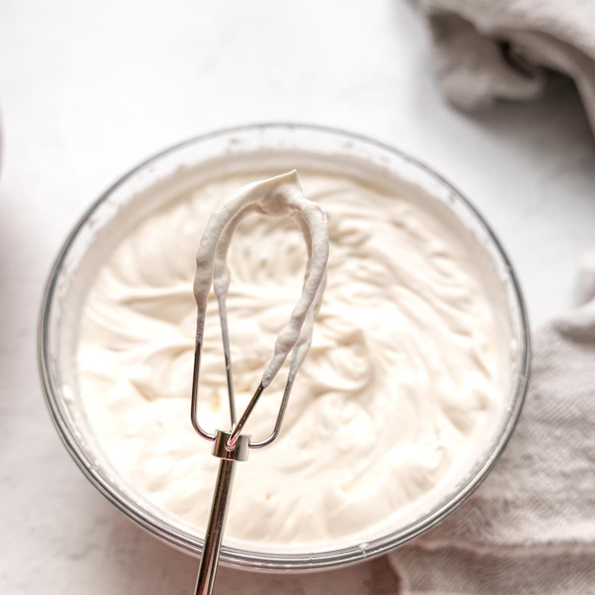 A metal whisk with stiff peaks of whipped cream held above a glass bowl filled with more whipped cream on a light surface.