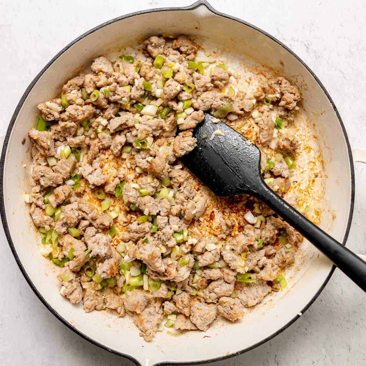 Ground meat and chopped green onions being cooked in a white skillet, stirred with a black spatula.