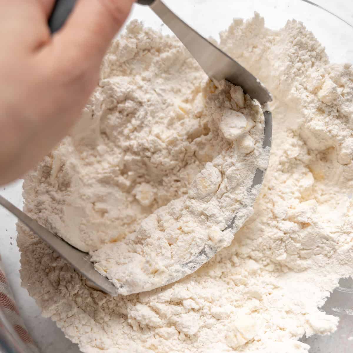A hand uses a pastry blender to cut butter into flour in a glass mixing bowl.