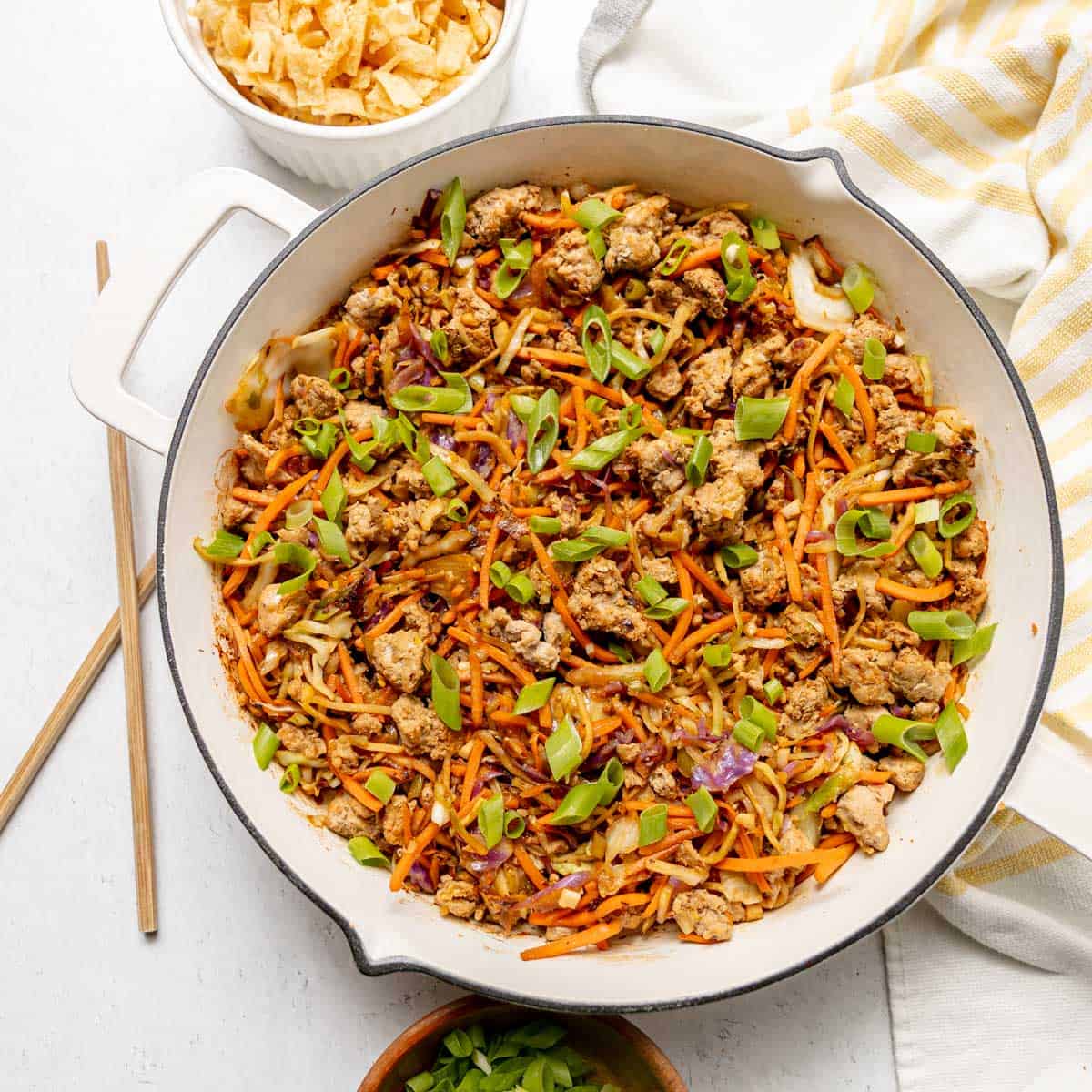 A skillet filled with ground meat, noodles, shredded carrots, chopped green onions, and cabbage, with a bowl of chips and a striped towel nearby.
