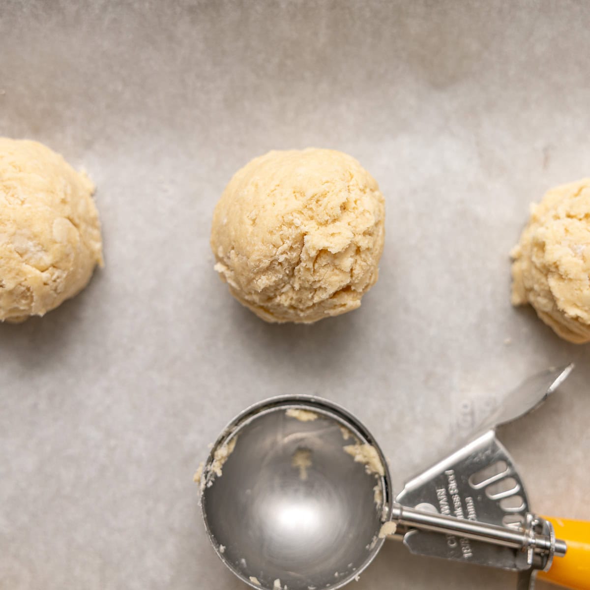Three scoops of raw cookie dough on parchment paper with a metal ice cream scoop beside them.