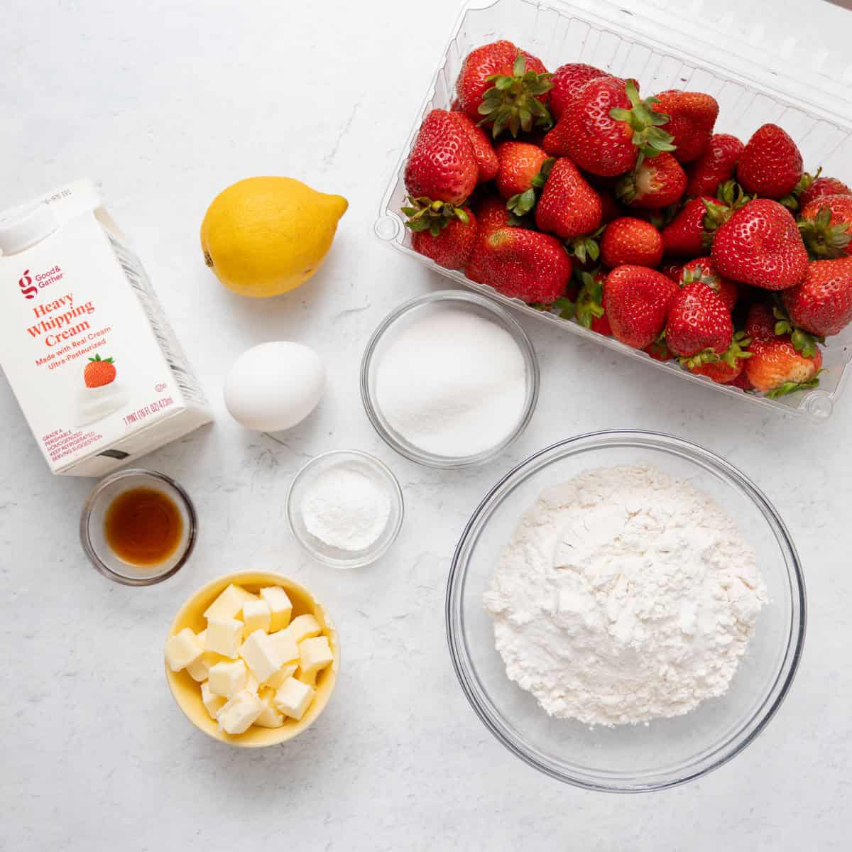 Ingredients for shortcakes: fresh strawberries, flour, sugar, an egg, vanilla extract, butter cubes, heavy whipping cream, and a lemon arranged on a white surface.