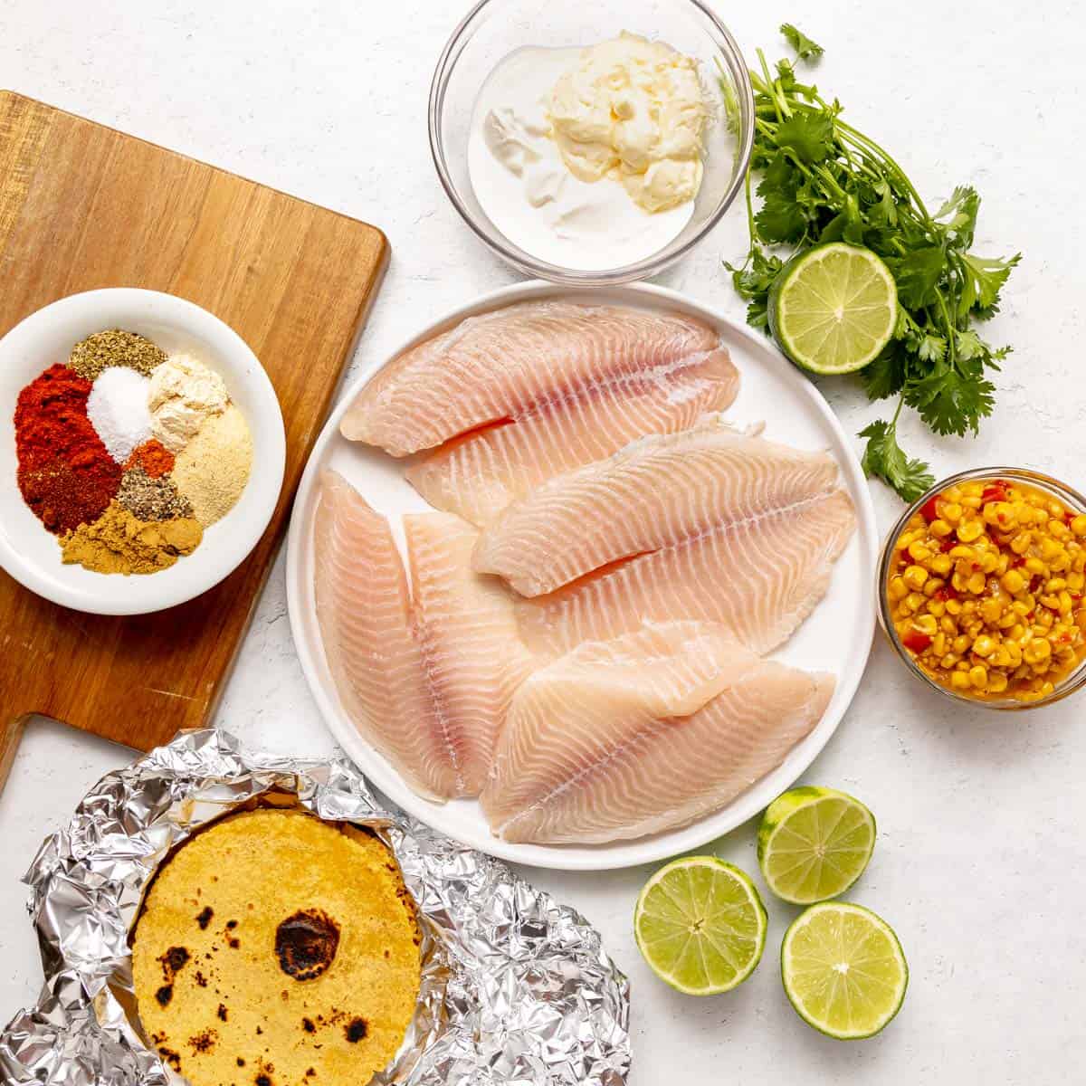 Raw fish fillets on a plate surrounded by bowls of spices, corn, mayonnaise, fresh cilantro, limes, and grilled tortillas on a cutting board.