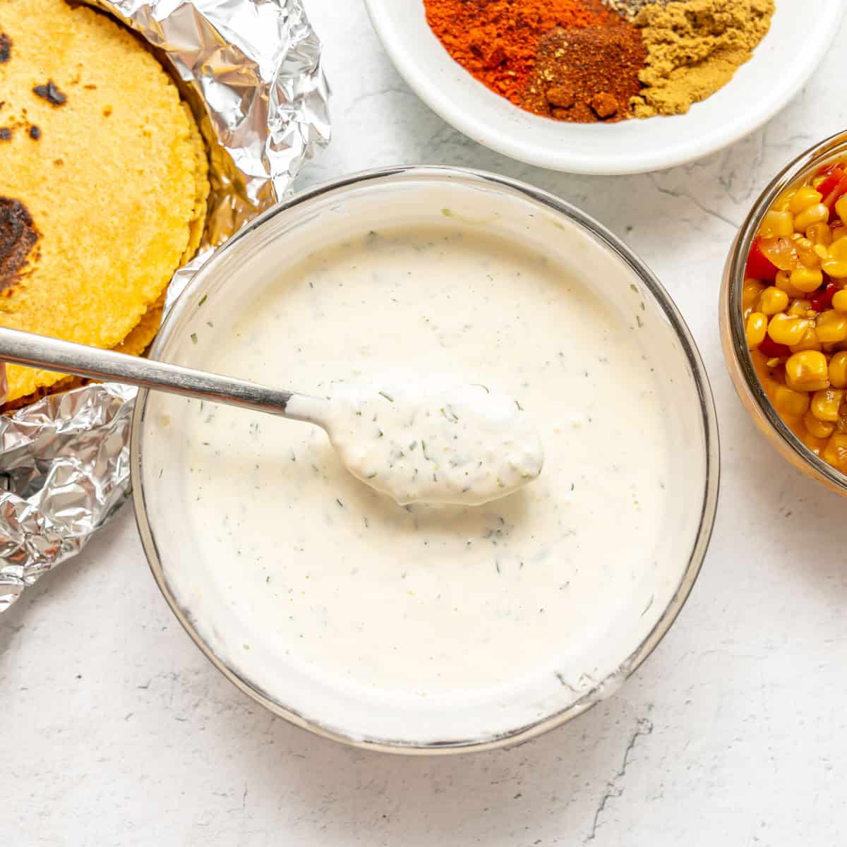 A bowl of creamy white sauce with herbs, a spoon inside, surrounded by tortillas in foil, a bowl of corn, and a small dish of spices.