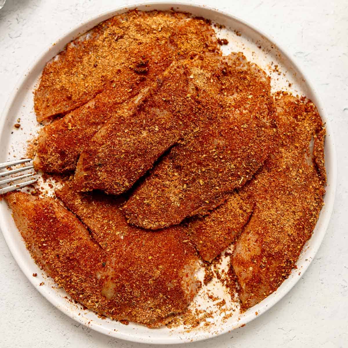 Raw fish fillets coated in a reddish-brown spice mixture are arranged on a white plate. A fork is partially visible on the left side of the plate.