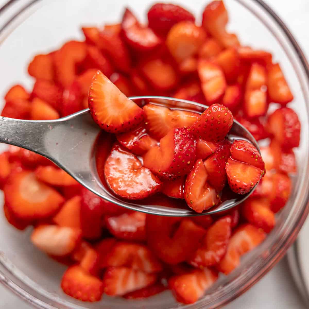 A metal spoon holds sliced strawberries over a glass bowl filled with more sliced strawberries.