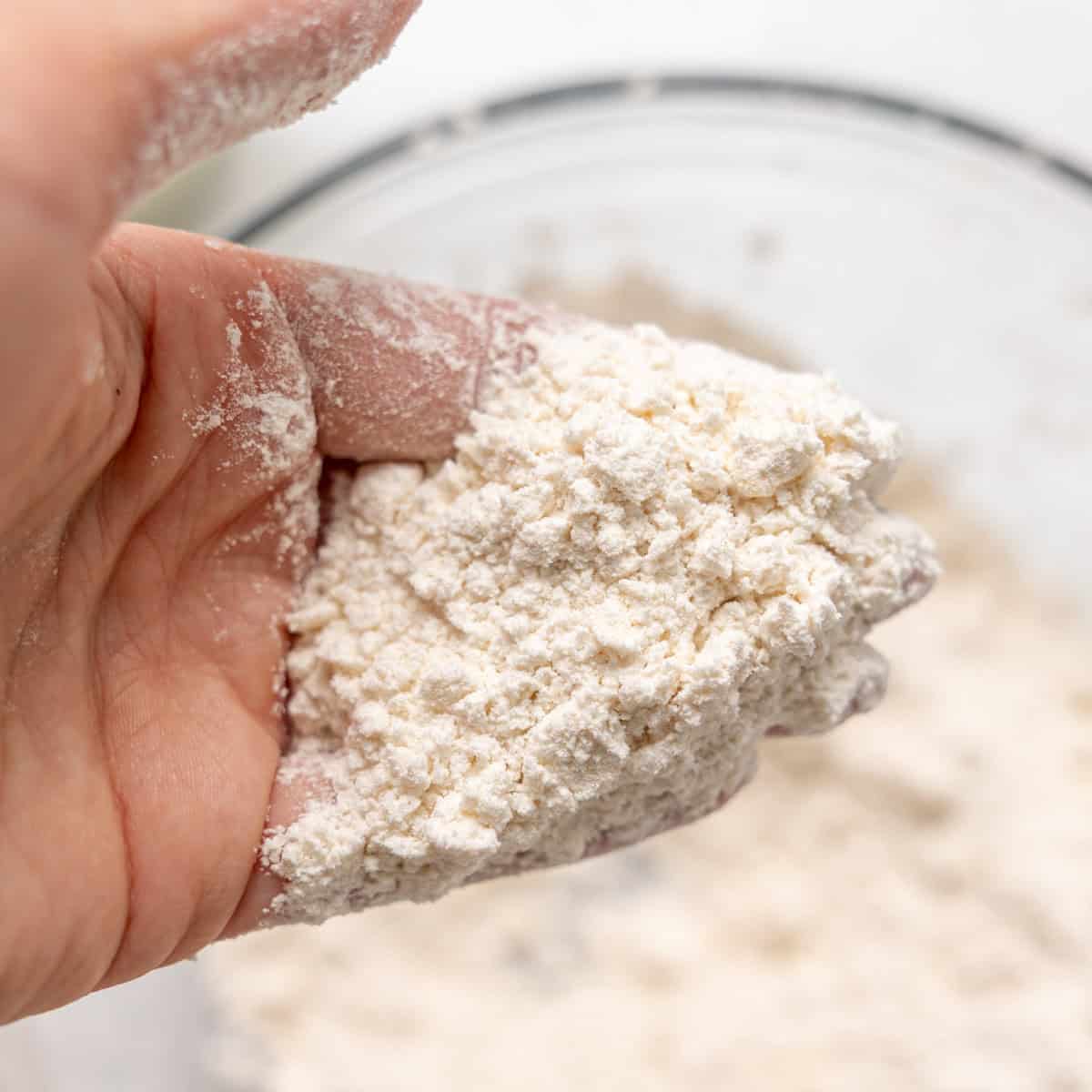 A hand holding a mixture of flour and small chunks of butter over a bowl, showing a crumbly texture.