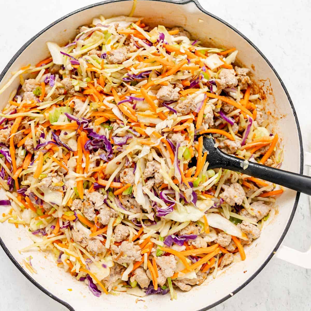 A skillet filled with cooked ground meat, shredded cabbage, carrots, and purple cabbage, with a black spatula resting in the mixture.