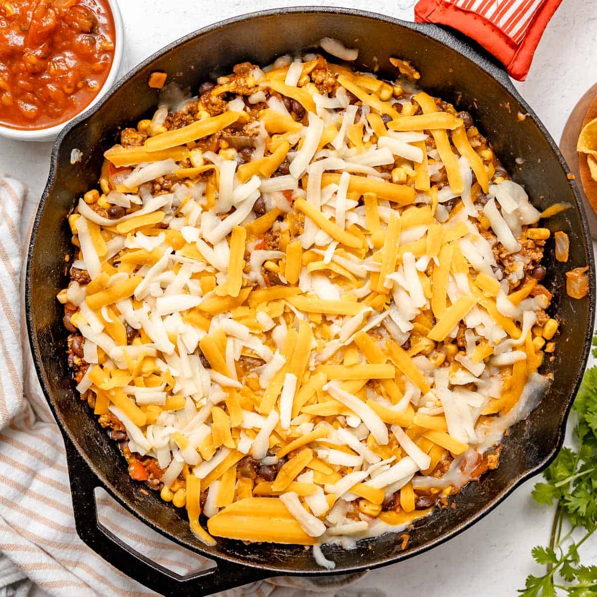 A cast iron skillet filled with a mixture of ground meat, beans, corn, and topped with shredded cheddar and mozzarella cheese.