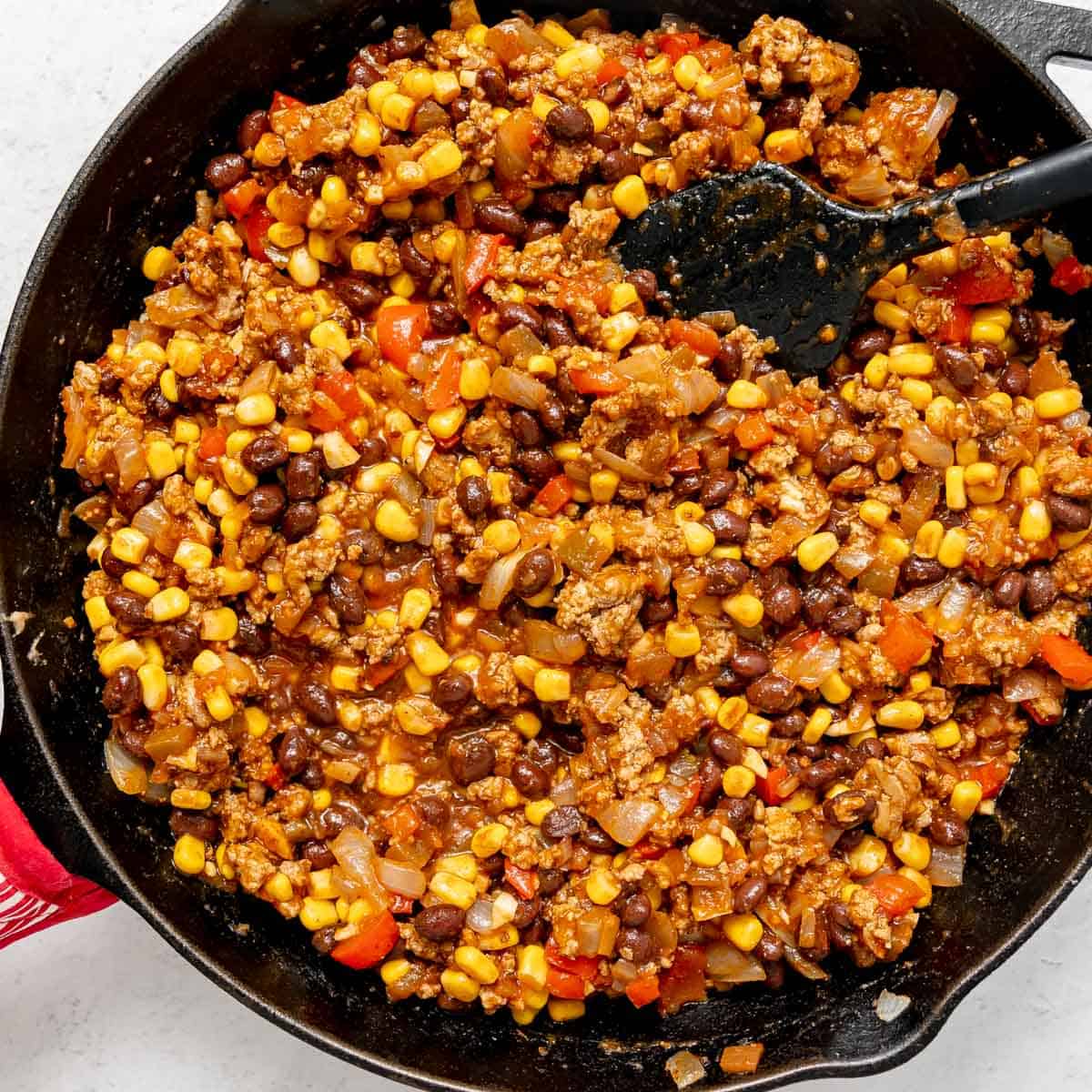 A skillet filled with a mixture of ground meat, black beans, corn, chopped onions, and red bell peppers, being stirred with a black spatula.